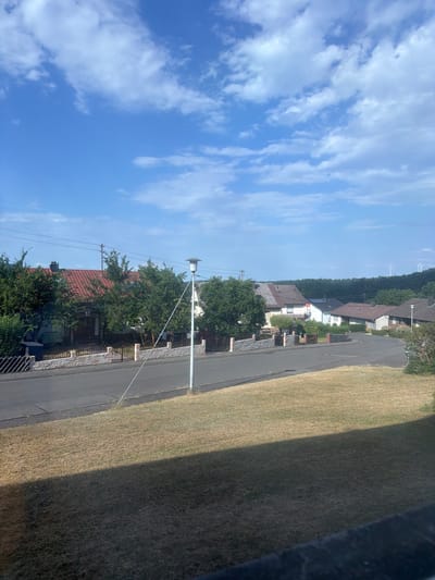 This media file shows a residential street in Idar-Oberstein, Germany, on a clear day. The scene is viewed from a slightly elevated perspective, looking down a gently curving paved road lined with houses and trees. The sky is a bright blue with scattered white clouds.
On the left side of the road, several houses with red-tiled roofs are visible, interspersed with lush green trees and gardens. Some houses have fences and low stone walls in front of them. A lamppost with a siren-like attachment stands by the roadside. Power lines are also visible, running along poles. In the distance, a forested hillside rises, with two white wind turbines visible on its crest.
On the right side of the road, a wide expanse of dry, light brown grass is present, casting a long shadow from an unseen object. The grass appears to be a lawn or a field.
The image appears to have been taken during the daytime, likely late morning or early afternoon, given the bright sunlight and the length and direction of the shadow. The weather is fair and sunny. No people or specific activities are depicted in the image. There is no visible text.