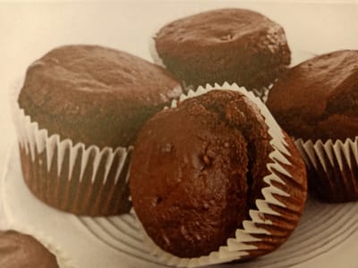The image displays a close-up of several chocolate cupcakes arranged on a light-colored surface. The cupcakes are in white paper liners, and their dark chocolate tops are visible. One cupcake in the foreground is slightly tilted. The background is out of focus and appears to be a plain, light color. There is no discernible urban environment or specific location identifiable within the image.