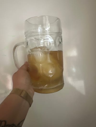 A hand wearing a gold mesh watch strap holds a large glass beer mug filled with a golden-colored liquid and ice. The mug is positioned in front of a plain, light-colored wall. There are faint green markings on the wall behind the mug. The scene is likely indoors. There is no information to determine the location in the city of Saint-Orens-de-Gameville, France.