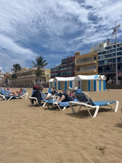 The image shows a sandy beach with people relaxing on lounge chairs. The scene is set in Las Palmas de Gran Canaria, Spain, at the coordinates 28.143, -15.433. In the background, there are buildings with balconies, palm trees, and a blue sky with white clouds. Three people are sitting on lounge chairs and reading. The scene is peaceful and relaxing, with the beachgoers enjoying the sunny day.
