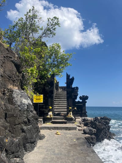 The image depicts a temple structure made of dark stone, possibly volcanic rock, situated on a rocky coastline. A paved pathway leads towards a set of stone stairs ascending to the temple entrance. The temple features ornate, traditional architecture with carved details. Lush green foliage grows on the rocks near the temple. A yellow sign with text in both Indonesian and English is posted on a metal stand to the left of the pathway, indicating attention and restrictions for entering the temple. The sea is visible in the background with waves crashing against the rocks. The sky is blue with scattered white clouds. The scene appears to be in a natural, coastal environment.