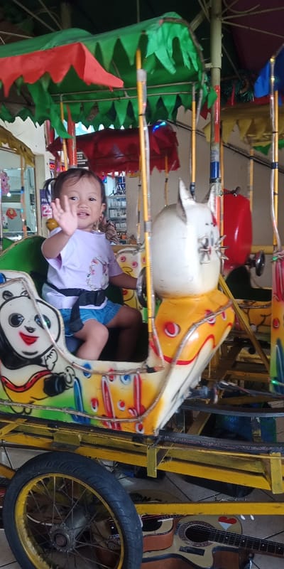 A young Asian child with dark hair in pigtails, wearing a light purple t-shirt and blue shorts, is seated in a colorful, panda-themed compartment of a small, mobile amusement ride. The child is secured by a black safety belt and is actively waving with their right hand, facing the viewer. The ride features a yellow metal frame, a prominent wheel with a yellow rim and black tire, and multiple animal-shaped seats, including a white cat-like figure to the child's right. Overhead, a multi-colored canopy, primarily green and red, shades the ride. The scene is located indoors within a commercial establishment in Cipanas, Indonesia, characterized by a tiled floor and a cluttered background displaying various merchandise on shelves, reflective surfaces, and signs. Two acoustic guitars, one featuring a red and white heart design resembling the Indonesian flag, rest on the floor below the ride's structure.
