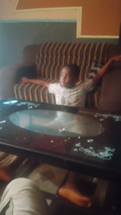 The image shows a young Black male sitting on a striped couch, with his arms outstretched. He is wearing a white t-shirt. In front of him is a dark, circular coffee table with a reflective surface. The background consists of a wall with a light-colored stripe at the top and a darker stripe below. The scene appears to be indoors, likely in a living room. The image quality is low, with a blurriness that makes specific details difficult to discern. There is no discernible information to place the scene in Asubi, Nigeria, or any specific city.