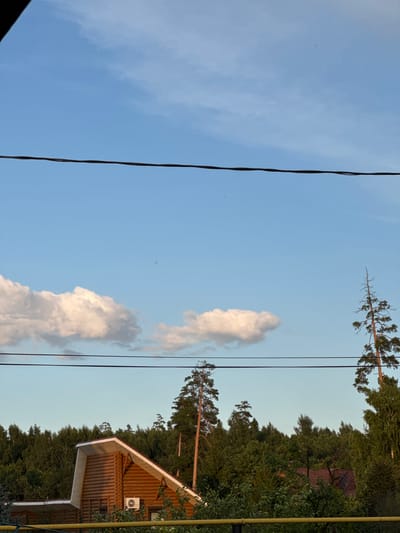 The image depicts a clear blue sky with scattered white clouds over a dense green forest. In the foreground, a wooden house with a prominent gable roof is visible on the left side. A small, white air conditioning unit is mounted on the exterior wall of the house. Power lines traverse horizontally across the sky. The scene appears to be a rural or suburban environment, possibly a dacha or country house in or near Белоус, Russia, given the architectural style and surrounding forest. There are no people or animals visible in the image. The overall impression is one of a tranquil summer day.