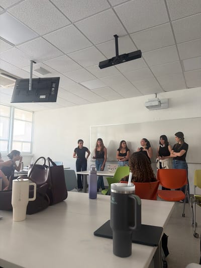 The image depicts an indoor scene within a modern classroom or conference room. The specific location within Monterrey, Mexico, is not discernible from visual cues.
The room features white walls and a white ceiling constructed of acoustic tiles, incorporating recessed rectangular LED light panels. A large window with multiple panes is visible on the left, providing natural illumination. Two flat-panel displays, showing their rear components, and one long, black overhead projector are suspended from the ceiling. A white, wall-mounted projector is positioned above a large whiteboard on the rear wall.
In the mid-ground, a group of six young adult females stands in a line facing forward, positioned in front of a white table and the whiteboard. They are uniformly dressed in dark tops and dark pants. From left to right: one female stands with hands on her hips; another holds a dark tablet or notebook; a third stands with arms crossed; a fourth gestures with both hands; a fifth holds an object in her left hand; and the sixth stands with arms crossed. Their posture suggests a presentation or group interaction.
In the foreground, a white table displays several objects. These include a dark grey Stanley tumbler with a handle and a straw, a white Hydro Flask mug with a handle, and a dark purple water bottle. A closed dark-colored tablet or laptop is positioned underneath the Stanley tumbler. A large, dark brown handbag is visible on a chair or table to the left. Partially visible seated individuals are present in the left and central foreground, with one individual in a green chair facing away from the camera, observing the standing group. Various chairs in colors such as orange, green, light blue, and yellow are arranged throughout the room.