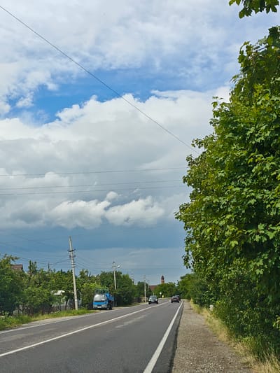 The image depicts a rural road scene in Deliatyn, Ukraine, under a partly cloudy sky with dramatic, dark gray clouds interspersed with patches of blue. A paved road with a white dividing line runs through the center of the frame, flanked by a gravel shoulder on the right and a grassy verge with dense green foliage. On the left side of the road, houses with red roofs and fences are visible behind a line of trees. A small blue truck is parked on the side of the road. Further down the road, several cars are traveling in the same direction. In the distance, a red-brick tower, possibly part of a church or historical building, rises above the trees. The right side of the image is dominated by a thick wall of lush green trees and bushes, with a power line visible in the upper portion of the frame. The overall impression is of a quiet, somewhat overcast day in a Ukrainian village.