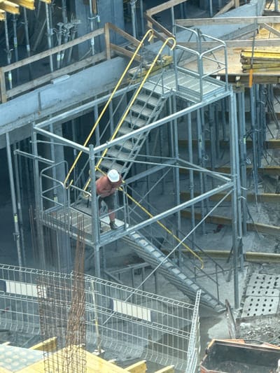 The image shows a construction site with a person wearing a white hard hat working on scaffolding. The person appears to be shirtless and is wearing shorts. The scaffolding is made of metal and has yellow railings. In the background, there are concrete structures, rebar, and other construction materials. The scene suggests ongoing construction work, possibly in Gdynia, Poland, given the context. The lighting and shadows suggest it is daytime. There are no visible signs of distress or specific emotions from the worker. The overall impression is a typical construction environment.