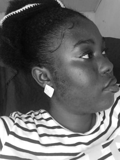 A black and white close-up image features a person with dark skin and hair pulled back, adorned with a white, knitted hair accessory. The individual wears a striped shirt and a square-shaped, textured earring. Their gaze is directed upwards and to the right. Facial features are prominent, including full lips, a defined nose, and a slightly visible area of hair growth on the cheek. The background is dark and somewhat indistinct.