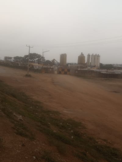 An outdoor, eye-level, medium shot captures a hazy industrial site in Sabon Gari, Nassarawa, Nigeria. The foreground is dominated by a dusty, reddish-brown dirt road that slopes down from left to right, with patches of sparse green grass growing on the embankment on the left. In the midground, a series of industrial structures rise against a pale, overcast sky. These include several tall, cylindrical silos, some appearing metallic and others concrete. To the left, a utility pole with wires is visible. Further left, trees with dense foliage are scattered. Several cranes, including a large tower crane, are present, suggesting ongoing construction or operation. Yellow and black checkered barriers are positioned near some of the structures. The background shows more buildings and greenery, blending into the misty atmosphere. The overall impression is one of an active industrial zone under a diffused, grey sky.
