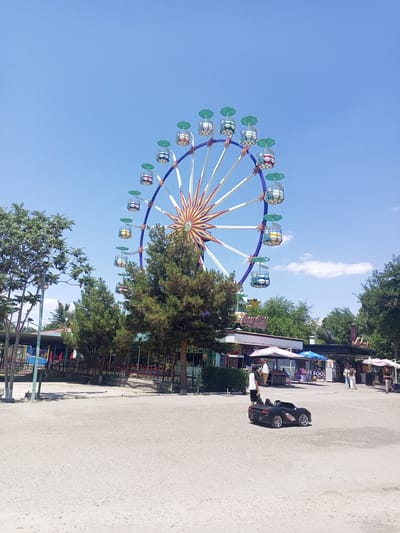 In a sun-drenched outdoor amusement park, likely located in Bukhara, Uzbekistan, a large Ferris wheel dominates the skyline against a clear blue sky. The Ferris wheel features colorful gondolas with teal-colored umbrellas. In the foreground, a black toy sports car is parked on a gravelly surface. The park appears to have several food stalls or kiosks, some adorned with umbrellas, and scattered trees provide shade. People can be seen walking in the background, some near the stalls and others further away. The overall scene suggests a typical day at a recreational facility.