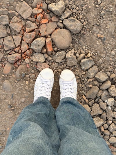 The image shows a downward view of a person standing on an outdoor ground surface composed of gravel, dirt, and broken rock fragments. The subject is wearing wide-leg, light-wash denim jeans and white sneakers with white laces. The location is inferred to be Rossiyskiy, Russia, based on the prompt, though there are no distinctive location markers visible within the image. No actions or interactions are depicted. The environment appears to be an unpaved area.