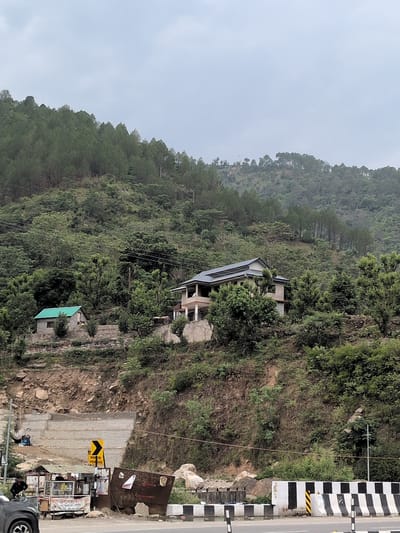 The image depicts a hillside scene in a mountainous region, likely in Sambal, India. A dense forest of pine trees covers the upper slopes of the hill. Below the forest, there are two houses situated on the hillside. The larger house, with a light-colored facade and a dark-tiled roof, is built on a terraced section of the slope. To the left and slightly lower down, a smaller house with a green roof is visible.
The foreground shows a roadside area. A road with black and white striped barriers runs horizontally across the bottom of the image. Adjacent to the road, there is a small roadside stall or shop with various items displayed. Several people are visible near the stall, including one person standing to the left and others in the background. A yellow road sign with a black arrow indicating a turn is also present. The ground in front of the stall appears to be a construction or work zone, with some debris and materials scattered around. A grey car is partially visible on the far left. The overall environment suggests a rural or semi-rural setting with evidence of development and daily life along a roadway. The sky is overcast, indicating a cloudy or possibly misty day.
