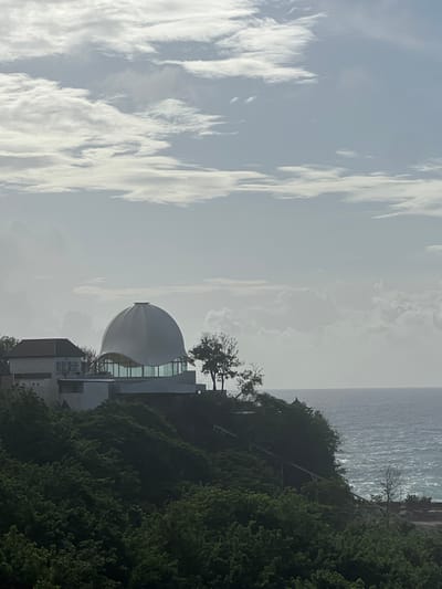 The image displays a panoramic view of the coastline in Uluwatu, Indonesia, under a partly cloudy sky. The primary subject is a distinctive modern building complex situated atop a verdant cliff on the right side of the frame. The central structure features a large, smooth, white, ovoid dome that appears to be the roof of a circular building. Beneath the dome, a section of the building has extensive curved glass windows, reflecting the sky. To the immediate left of this domed structure, a portion of another building with a traditional tiled or gabled roof and light-colored walls is visible.
The buildings are surrounded by dense, dark green foliage covering the cliff face. A few sparse, deciduous-looking trees are visible near the domed building and further down the cliff. On the right side of the image, the blue-gray ocean stretches to the horizon, with visible whitecaps indicating active waves near the shore. A faint, low-lying structure or rock formation is discernible at the water's edge on the far right. The sky occupies approximately the upper third of the image, displaying a mix of bright white cumulus and stratiform clouds against a lighter blue-gray haze. No individuals or specific actions are visible in the scene.