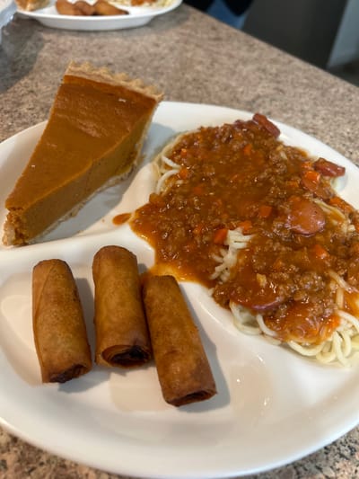 The image displays a white, partitioned plate containing a meal, resting on a light-colored, granular countertop. In the upper-left partition, there is a single slice of pumpkin pie with an orange custard-like filling and a light-brown crust. In the upper-right partition, a serving of spaghetti is visible, covered in a reddish-brown meat sauce that includes ground meat, diced orange vegetables (likely carrots), and distinct slices of what appear to be hot dogs. The lower-left partition holds three golden-brown, fried cylindrical spring rolls, arranged in a row, with some filling visible at the ends of two rolls. In the blurred background, another white plate containing indistinguishable food items and a dark fabric object are partially visible. No people, specific actions, or interactions are discernible in the foreground. The specific location within Thousand Oaks, United States, cannot be determined from the visual information present in this image.