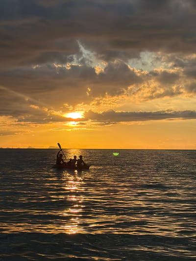 A silhouette of four people in a kayak on the water against a sunset in Baan Tai, Thailand. The sun is setting on the horizon, casting a golden glow across the sky and reflecting on the rippling water. The clouds in the sky are dark and dramatic, with streaks of orange and yellow from the setting sun. The kayak is positioned in the middle of the frame, with one person at the front holding a paddle, and three others seated behind. In the distance, faint outlines of islands can be seen.