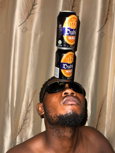 A man wearing sunglasses balances two Dubi Malt cans on his face. The cans, lined up in a row, rest on his eyes, supported by the sunglasses. The cans feature a gold crown design and product information in black and white text. The man has dark skin, a beard, and exposed shoulders. The background consists of a shiny, textured beige fabric with a feathery pattern. The image is oriented horizontally.