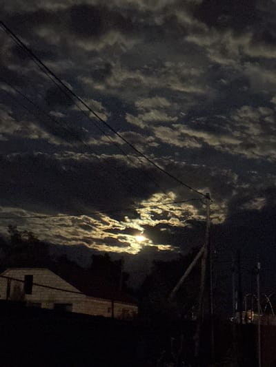 The image depicts a night scene with a full moon partially obscured by clouds. The moon's light illuminates the clouds from behind, creating a dramatic effect. In the foreground, there is a low-lying building, possibly a house, with a dark roof and a single visible door. Several power lines crisscross the sky. The environment appears to be somewhat rural or suburban, with trees visible on the left side and a wooden pole on the right. The overall atmosphere is dark and somewhat eerie due to the lighting conditions. The location is Ləcət, Azerbaijan.