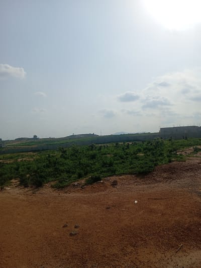 The image depicts an outdoor scene in Karsana 2, Nigeria, characterized by a construction site and surrounding undeveloped land. The foreground consists of reddish-brown, dry earth with scattered small stones and pebbles. A white plastic bag and a thin stick are visible on the ground in the mid-foreground.
In the mid-ground, a densely vegetated area with green shrubs and plants covers the landscape. This vegetation extends towards a low, stone-walled perimeter that encloses the area. Beyond the wall, the terrain rises into gently sloping hills.
The background features a large, multi-story building under construction, made of grey concrete blocks, with exposed rebar at the top. Further in the distance, other buildings and structures are visible, along with more hills and mountains on the horizon. The sky is overcast with scattered clouds, and the sun is faintly visible, creating a bright, washed-out effect in the upper right portion of the image. The overall impression is one of ongoing development in a semi-rural or exurban setting. There are no people or animals visible in the frame.