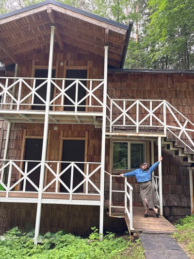 A young woman stands on a wooden ramp in front of a rustic, wooden cabin. She is wearing a blue denim jacket and a long leopard print skirt, and her arms are outstretched. The cabin has multiple levels with white railings and wooden siding. There is lush green foliage surrounding the cabin, with trees visible in the background. The setting appears to be a natural environment, likely in a rural area.