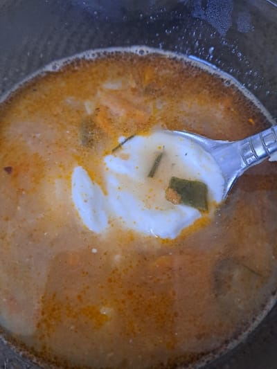 The image displays a close-up, top-down view of a reddish-orange liquid dish within a dark-rimmed container, located in the city of Bel'kovo, Russia. The surface of the liquid exhibits numerous small, orange fat droplets and a distinct frothy, dark-orange ring along the inner edge of the container. A metallic spoon is partially submerged in the liquid, holding a white, viscous substance, consistent with dairy, along with several dark green fragments and a smaller, light-orange food particle. Additional light-colored and dark-green food particles are observed suspended within the main body of the liquid. The spoon's presence indicates an action of serving or consumption.