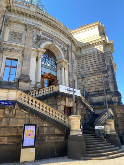 The image shows the exterior of a building in Dresden, Germany. The building is made of stone and features ornate architectural details, including arched windows, columns, and sculptures. The name "OKTOGON" is visible above a large window.
Signs are present near the entrance, with one indicating "OPEN" and another advertising an event called "ALLES SO SCHÖN BUNT HIER!". A street sign labeled "uenkirche" is also visible, indicating the location is near a church. The photograph is taken from a low angle, with a clear blue sky in the background.