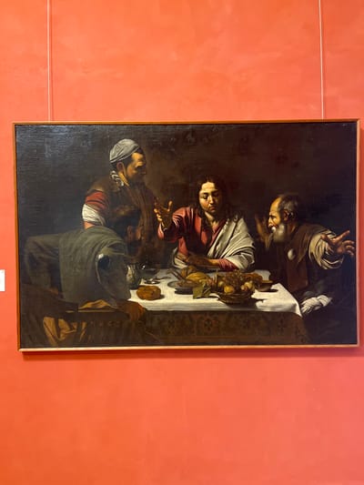 The painting depicts a scene of a shared meal, likely a religious subject given the common portrayal of Jesus at a table. Four figures are present. On the left, a man in a grey cap and tunic leans over the table, his back to the viewer. To his right, a central figure, possibly Jesus, is dressed in a red garment over a white robe, with his head tilted down and his right hand gesturing. Across the table, an older man with a beard and brown tunic faces the central figure, his mouth open as if speaking, and his arms extended. Behind the man in the grey cap, another figure in a brown and red garment with a ruffled collar looks towards the center. The table is covered with a patterned tablecloth and laden with food, including bread, a basket of fruit (grapes and other items), and various vessels. The background is dark, creating a dramatic contrast with the illuminated figures and table. The painting is framed by a simple wooden border and is hung on a vibrant coral-pink wall. The image does not provide any information about the specific location in Palermo, Italy.