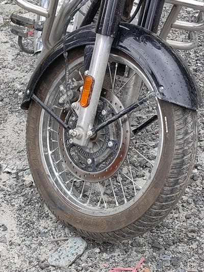 The image shows the front wheel of a motorcycle parked on a gravel surface. The motorcycle has a black fender and a silver suspension. The tire is a MRF Zapper. There is a brake disc visible on the wheel. The ground is covered with loose gravel and some debris. The scene appears to be outdoors, in a place that may be near a construction site or a rural area. The specific location within Solang, India, cannot be determined from the image alone. There are no people visible in the image.