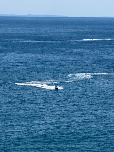 An aerial view captures a vast expanse of the deep blue Adriatic Sea with a distant, hazy coastline of mountains and landforms. In the foreground, a person rides a jet ski, creating a prominent white wake that curves across the water. Further in the distance, another jet ski with two riders is visible, also leaving a smaller white trail. The sea is moderately choppy with a textured surface. The sky above is a pale, hazy blue, with a thin line of clouds near the horizon.