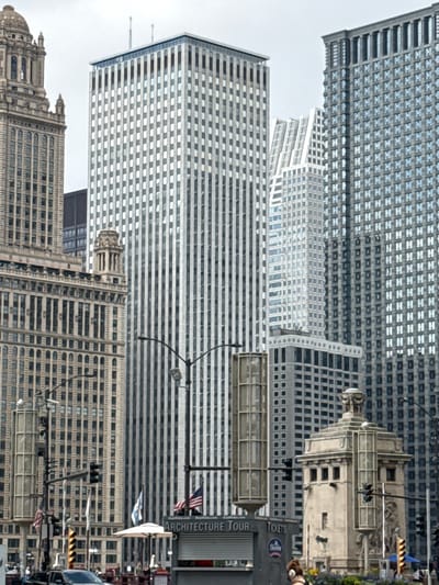 This media file captures a view of several tall buildings in an urban setting, likely Chicago. The architecture varies, with some buildings featuring a repetitive grid of windows and others exhibiting more ornate, classical designs, including domes and intricate detailing. The perspective is angled upwards, emphasizing the height of the structures. The sky appears overcast, suggesting a cloudy or hazy day.
Among the buildings, there are various objects visible. Traffic lights are present, indicating a street-level view or an intersection. Flags, including American flags, are displayed on some buildings. There are also signs, one of which reads "ARCHITECTURE TOUR" and another with "TICKETS" and a "Sears" logo, suggesting a tour bus or information kiosk. The presence of cars and what appears to be a tour bus hints at an active tourist area. Though not the primary focus, there are glimpses of people in the background, and a person's head with brown hair is visible in the lower right corner, possibly taking a photo or observing the scene.
The overall impression is of a bustling city environment, rich in architectural history and offering attractions for visitors. The time of day is not definitively clear due to the overcast sky, but it appears to be daytime.