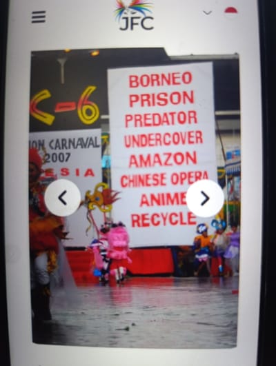 The image shows a street scene in Kecamatan Arjasa, Indonesia, with several people in colorful costumes walking away from the camera in front of a sign that reads, "BORNEO PRISON PREDATOR UNDERCOVER AMAZON CHINESE OPERA ANIME RECYCLE". The people are wearing brightly colored clothes and some have masks. There are arrows on the screen, and the words "C-6", "ION CARNAVAL 2007", and "ESIA" are also visible. The ground is wet, and the scene is a street or a public space. The image is likely taken during a carnival or parade. The image suggests that the scene is a festive one, with people enjoying themselves. There is a sense of movement and energy in the image. The image is somewhat blurry. There are two arrows on the screen that suggest this is a page in an app or a website. The image was likely taken from a phone or tablet.