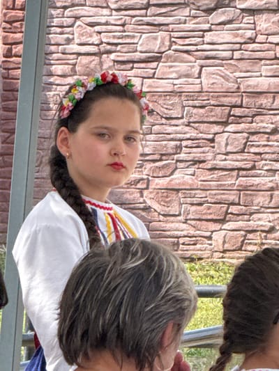 The image shows two people in an outdoor setting with a brick wall in the background. One person, a young girl, is in the foreground, wearing a white shirt with colorful embroidered trim and a flower crown on her head. She has dark hair braided and is looking towards the camera with a neutral expression. The other person is partially visible behind her, with dark hair. The scene appears to be a casual gathering or event, possibly a festival or a cultural celebration given the traditional-looking embroidery on the girl's shirt and her flower crown. The lighting suggests it might be daytime, with bright, natural light. There is no visible text in the image. The location context provided is Pordim, Bulgaria.