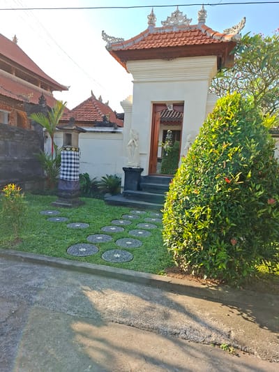 The image depicts the entrance to a building in Blahbatuh, Indonesia, with traditional Balinese architectural elements. A pathway made of circular stone pavers is laid across a green lawn. To the left, a stone lantern is adorned with a black and white checkered cloth. In front of the entrance, two white statues stand on pedestals. A large, lush green shrub occupies the right side of the frame. The building features a red-tiled roof with ornate decorative elements. The sky is clear and blue. The scene appears to be a residential or temple entrance, bathed in sunlight.