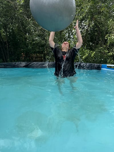 A person is in a swimming pool in a yard in Khokhryaki, Russia, holding a large, gray exercise ball above their head. The person is wearing a wet, black t-shirt, and water is splashing around them. The pool is filled with clear blue water, and the edge of the pool is lined with black plastic. Trees and greenery are visible in the background, suggesting an outdoor, residential setting.
