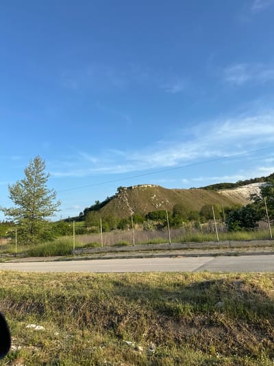 The image depicts a landscape in Beloslav, Bulgaria. A road runs along the right side of the frame, bordered by a concrete barrier and grassy verge. Parallel to the road is a hillside covered in vegetation. The top of the hill shows a cliff edge. A single tree is in the lower left of the image, while power lines extend across the sky. The sky is clear blue with some wispy white clouds.