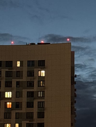 The image captures a nighttime view of a modern apartment building in Pushkino, Russia. The building's facade is predominantly a light beige color, with a grid-like pattern of windows. Several windows are illuminated from within, casting warm yellow and orange light onto the dark sky. The top of the building features a darker, notched section, possibly housing mechanical equipment or decorative elements.

Against the dark, overcast sky, three red lights are visible, spaced vertically along the left edge of the frame. These lights appear to be part of an aviation warning system, indicating the height of a nearby structure, likely the building itself or an adjacent one. The sky is a deep, moody blue-gray, with hints of lighter, cloud-like formations that are indistinct in the low light. The overall impression is one of an urban environment at dusk or night, with the glowing windows providing points of life and warmth against the imposing structure and the darkening sky. The weather appears to be cloudy, contributing to the dim lighting conditions. No people are visible in the image, and there is no discernible text. The scene evokes a sense of quiet solitude, typical of a city after dark.