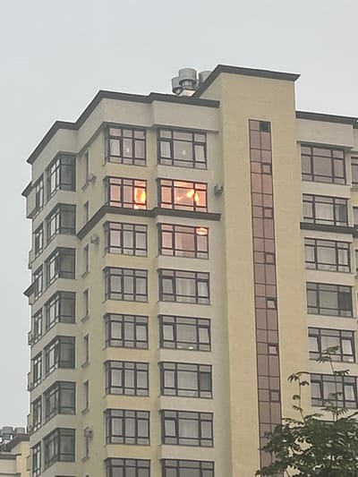 The image shows the facade of a multi-story residential building in Kemerovo, Russia. The building is light yellow with dark brown window frames. Several windows are lit from within, suggesting occupancy. A narrow vertical section of the building features tinted glass panes. The sky is overcast. A tree with green foliage is partially visible in the lower right corner of the frame.