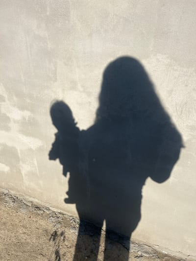 The image displays a shadow of a person on a textured, light-colored surface, likely concrete or stucco. The shadow is dark and sharply defined, suggesting bright, direct sunlight. The figure in the shadow appears to be wearing clothing, with a discernible head and torso, and possibly legs. The overall impression is that of a person standing or walking outdoors. The surface has a rough, granular texture with some variations in color and smoothness. In the lower right portion of the image, a transition to a rougher, uneven surface is visible, with cracks and debris, suggesting an outdoor environment. The light and shadow indicate it is likely daytime. No text is visible in the image.