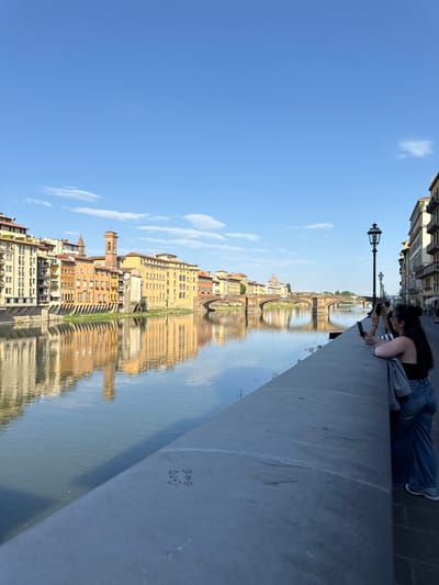 The image captures a picturesque view of the Arno River in Florence, Italy, with the Ponte Vecchio visible in the distance. The riverfront is lined with historic buildings showcasing typical Florentine architecture, featuring pastel-colored facades and balconies. The clear blue sky is dotted with a few wispy clouds, reflecting beautifully on the calm water. On the right side of the frame, two young women are standing by the river wall, engrossed in taking photos with their smartphones. The woman in the foreground is leaning on the wall, wearing a black top and wide-leg jeans, with a grey jacket draped over her shoulder. The other woman, partially visible, also appears to be taking a photo. The river wall itself has some faint graffiti markings. The scene evokes a sense of tranquility and tourism, characteristic of Florence's iconic cityscape.