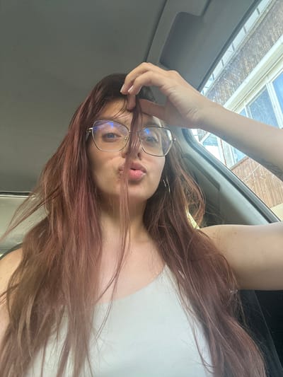A young woman with long, reddish-brown hair and glasses is in the driver's seat of a vehicle, looking towards the camera. She is making a puckered lips gesture, and her right hand is resting on top of her head, touching her hair. The interior of the car is visible, including the dashboard and a window showing a building with multiple windows. The image appears to be taken from a low angle, looking up at the woman. The setting is not specific to Tryavna, Bulgaria.