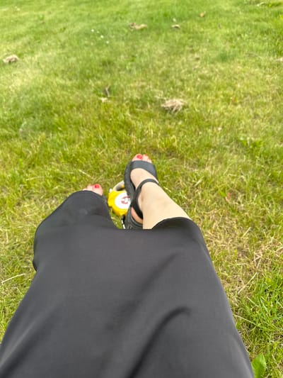 The image shows a person's legs and feet, wearing black sandals and a black skirt, sitting on a grassy area. A yellow object with a red accent is visible near the person's feet. The background is out of focus green grass. The scene appears to be an outdoor public space, possibly a park. Riihimäki, Finland is a city known for its parks and green spaces, making it a plausible location for such a scene.