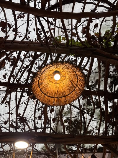 The image depicts an interior scene in Bencongan, Indonesia. A light bulb encased in a circular, straw-like lampshade is the focal point, suspended against a background of a light-colored, possibly translucent material, possibly a ceiling, which is overlaid with a dense network of dark, woody branches and vines. The branches appear to be part of a decorative structure. A second, smaller, round light fixture is visible on the lower right.