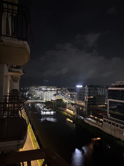 A nighttime view from a balcony shows a cityscape in Pasay, Philippines. A dark, reflective body of water, likely a canal or river, runs through the scene. Buildings with illuminated windows and striking architectural designs line the waterfront. One building features a unique, honeycomb-like facade, while another displays the "FLEXPLEX" logo illuminated in white. Construction cranes are visible in the distance, indicating ongoing development. The sky is dark and cloudy, with some lights from the city reflecting on the water's surface. The foreground shows parts of a balcony with railings and a worn concrete railing.