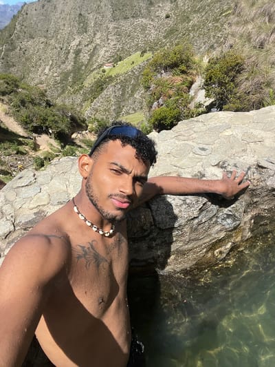 A shirtless man with curly hair and sunglasses on his head leans against a rock, his right arm resting on the rock's surface. He is looking towards the camera with a slight smile. He wears a necklace made of shells. The man has tattoos on his chest and shoulder. The background shows a mountainous landscape with green vegetation and a small house on a hillside. The man is in a body of water, and the rock he is leaning on is partially submerged. The image appears to have been taken outdoors during the daytime. The location is described as being in Mucurubá, Venezuela.