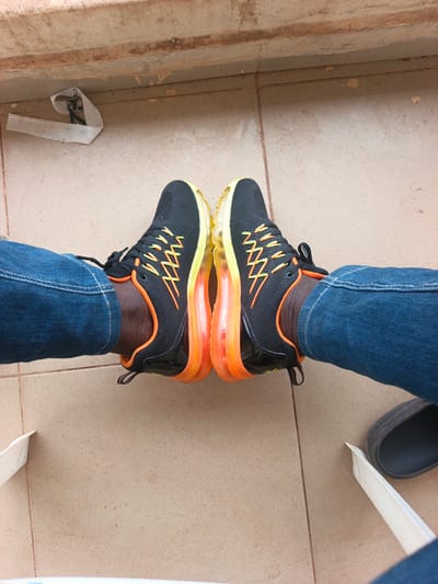 The image shows a person's legs from the knees down, wearing blue denim jeans and black athletic shoes with orange and yellow accents. The shoes have a thick, transparent orange and yellow sole that appears to be an air cushion. The person is sitting on a tiled floor with their legs extended forward. To the left of the frame, a small piece of white paper is crumpled on the floor. In the background, there is a ledge or step made of reddish-brown material. To the right of the frame, a dark-colored slip-on shoe or sandal is partially visible on the floor. The lighting suggests it is daytime, possibly outdoors given the natural light and the tiled floor. There is no visible text in the image.