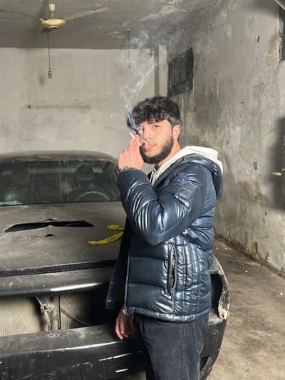 A young man with curly hair and a beard stands in what appears to be a garage, leaning on a damaged black car. He wears a shiny navy puffer jacket over a white hoodie and dark pants. Smoke curls upwards from a cigarette held in his hand. The garage has a concrete floor and walls, with a vintage-style ceiling fan. The overall atmosphere suggests a somewhat gritty or industrial setting.