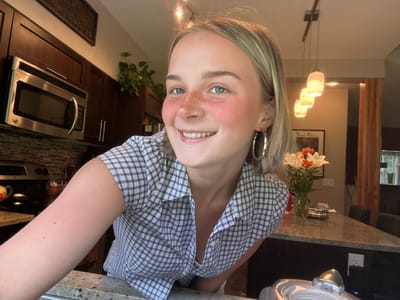 A woman is shown in an indoors setting. She is wearing a blue and white checkered shirt and large hoop earrings. Her face is the focal point; she has freckles across her nose and cheeks and her hair is blonde. To her left is a bouquet of flowers in a vase on a granite countertop. Behind her is a modern kitchen with dark wooden cabinets and a stainless-steel microwave. Pendant lights hang in the background. The location is inside an indoor space and there is no clear indication of the city, Whistler, Canada.