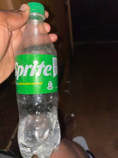 A person is holding a plastic bottle of Sprite in their hand. The bottle is mostly empty and has a green cap and a green label. The background is dark and indistinct. There is no specific information to indicate this image was taken in Kurunduma, Nigeria.