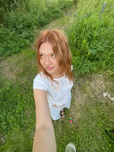 A young woman with long, reddish-brown hair lies on her back in a grassy field. She is wearing a white t-shirt with a pattern and grey pants. Her left arm is extended towards the camera, and she is looking up, smiling with her eyes wide. Her right leg is bent, and she is wearing a brown sandal.
The setting is a natural outdoor environment, likely a field or meadow, with lush green grass and some wildflowers. In the background, there is more vegetation, including tall grasses and some green plants with leaves. On the ground near her right foot, there is a green and red object that appears to be a firecracker or a similar novelty item, and a clear plastic cup.
The image appears to be taken during the daytime, possibly late afternoon or early evening, given the soft lighting. The weather seems to be pleasant, as the woman is outdoors in light clothing. There is no explicit indication of a specific event or activity taking place, but the relaxed pose and smile suggest a casual and enjoyable moment.
The text on the woman's t-shirt is partially visible and appears to include the word "DIVINE" in a decorative font.
The image has a natural, candid feel, possibly a selfie taken by the woman herself or by someone with her. The orientation of the photo, with the woman's head at the top and her feet towards the bottom, is unusual and suggests she might be lying down while taking the picture.