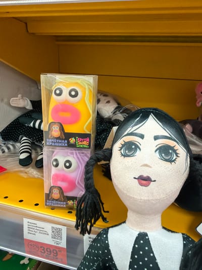 This media file presents a close-up view of merchandise within a retail setting, likely a toy aisle in a store located in Berezniki, Russia. The primary subject is a soft-stuffed doll, positioned horizontally, with a pale face, striking large eyes, small red lips, and black hair braided into pigtails. The doll is dressed in a black and white polka-dotted outfit, partially visible. Its overall aesthetic, characterized by its pale complexion and dark features, suggests a gothic or quirky style.

In front of the doll, two smaller, rectangular packaged items are displayed. These appear to be stress toys or squishies, one in yellow and the other in purple, each featuring large cartoon eyes and a pink "0" or "O" design. The packaging displays Russian text, including "Крутой ЗАМЕС АНТИСТРЕСС" (Cool MIX ANTISTRESS) and "ЗАЧЁТНАЯ КРАШИХА" (Cool CRUSH/BEAUTY).

To the right of the items, a retail price tag is visible. It identifies one of the items (likely the large doll) as "Игрушка мягконабивная BIGGA Аксолотль, 28см" (Toy soft-stuffed BIGGA Axolotl, 28cm) from China. The price tag shows a discounted price of 399.00 Russian Rubles, marked down by 45% from an original price of 736 Rubles. The background consists of solid yellow shelving, typical of a store interior, illuminated by artificial lighting. No individuals are present, and the scene focuses entirely on the products on display.