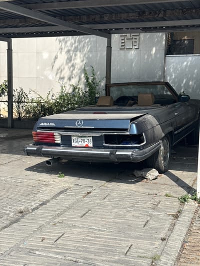 A blue Mercedes-Benz 380 SL convertible is parked under a metal structure, possibly a carport, in what appears to be a residential area in San Pedro Garza García, Mexico, as indicated by the Tamaulipas license plate "XGT-28-36." The car shows signs of disuse, covered with dust or dirt. The left door is ajar. Plants grow near the vehicle and the adjacent building wall. The ground is paved with concrete.